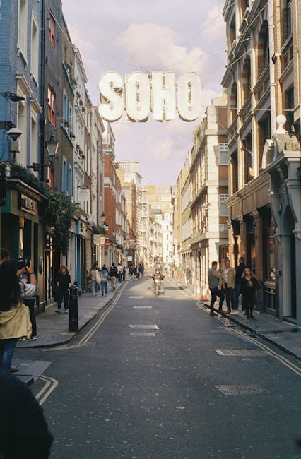 A busy city street in Soho, featuring a mix of historic and modern buildings with ornate facades and large windows. Pedestrians are walking along the sidewalks, some browsing shop windows or engaging